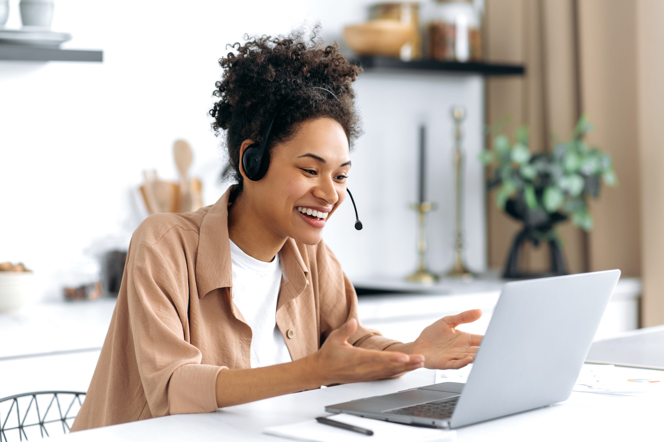 Friendly pretty, african american girl with headset, freelancer, designer, student sits at desk in the kitchen, talks via video communication, work or study remotely, gesturing with her hands, smiling